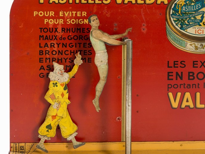The acrobat comes over the bar backwards and releases for a one-hand grip. His clown buddy enthuses for the viewers.